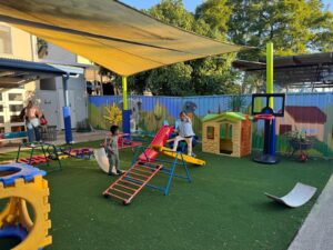 Outdoor play area at daycare centre in Seven Hills featuring children enjoying slides, climbing structures, and playhouses under shaded supervision.
